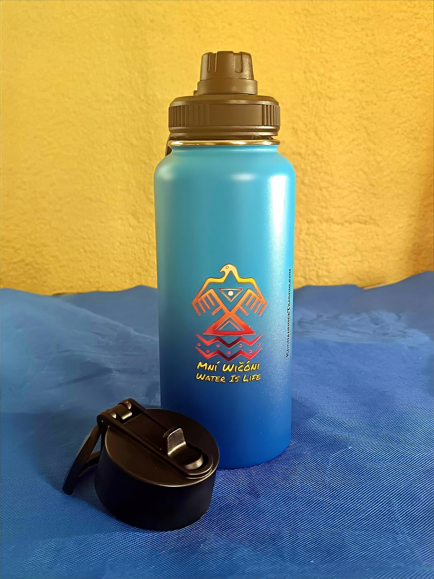 Kinnikinnick Trading Co. Water Bottle Blue water bottle with black cap and strap on blue surface against yellow wall.